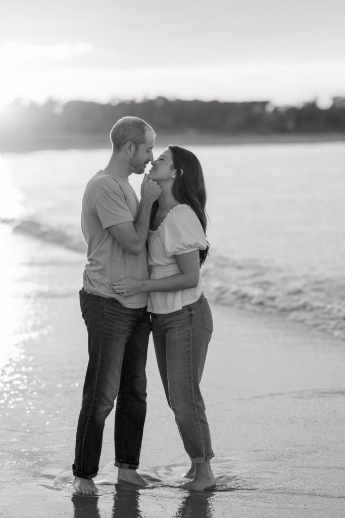 Black-and-white portrait of a couple standing barefoot in the ocean during sunset while waves roll around them, inspired by timeless coastal engagement photo locations in Massachusetts.