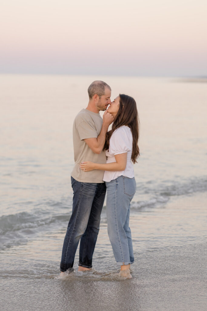 Couple kisses barefoot in the shallow water beneath pastel sunset skies during a romantic coastal engagement session, reflecting the airy feel often associated with Crane Beach engagement photos.