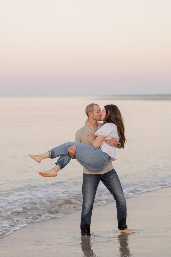 Man lifts his partner in shallow ocean water beneath a pastel sunset sky during a playful coastal engagement session, inspired by romantic beach engagement photos Massachusetts couples often search for.