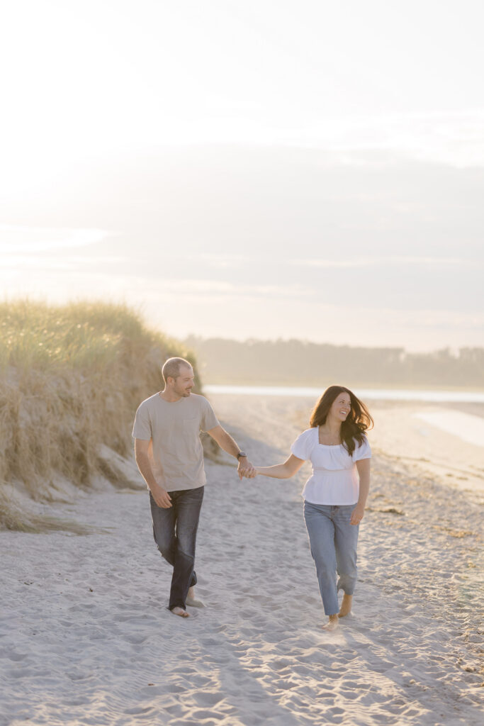 Couple walks barefoot beside tall beach grass with warm evening light filtering across the sand dunes, photographed at one of the more scenic engagement photo locations Massachusetts offers.