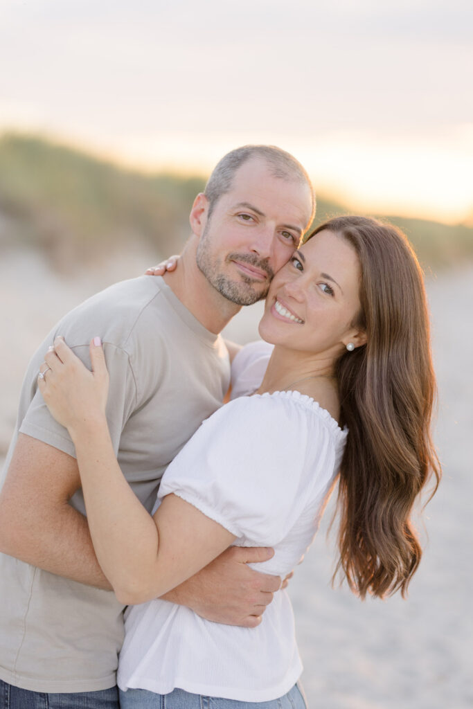 Close-up portrait of a couple embracing on the beach with warm sunset tones and soft dunes behind them, reflecting the relaxed atmosphere often seen in engagement photos Massachusetts sessions.