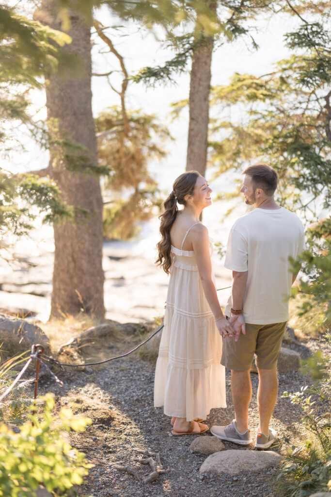 Couple stands together beneath tall trees overlooking the water during golden hour portraits, photographed with the soft editorial feel many Massachusetts engagement photographers are known for.