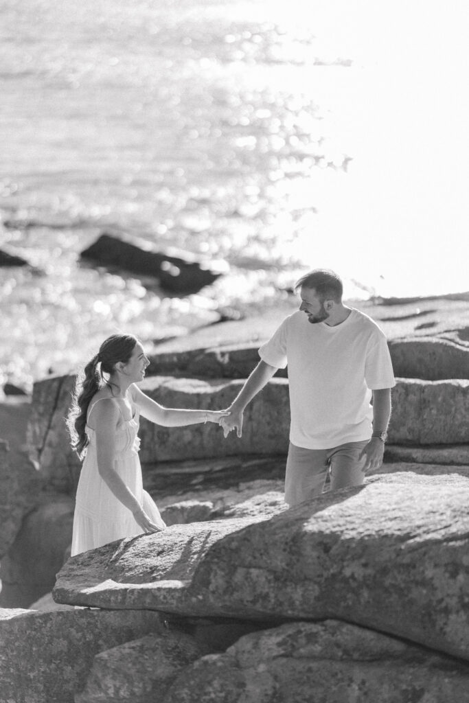 Black-and-white portrait of a couple climbing across seaside rocks with shimmering water behind them during a documentary-style coastal session, inspired by destination engagement photography near Massachusetts and Rhode Island.